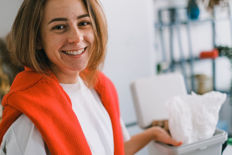 Positive young female with colorful sweater on shoulders smiling and looking at camera while throwing rubbish into rubbish bin in light kitchen at home